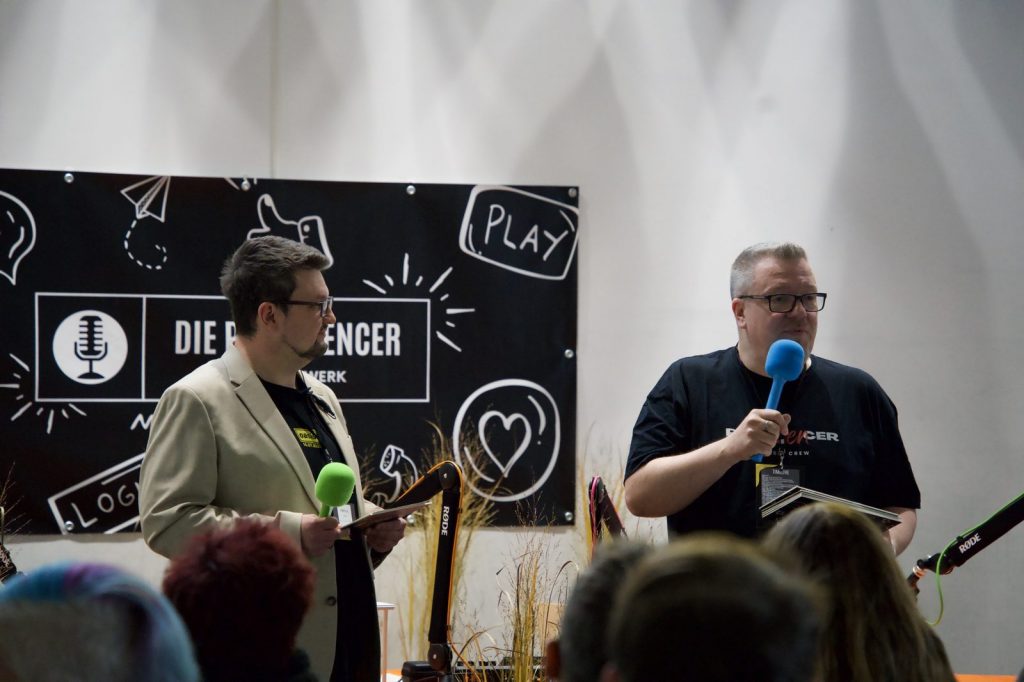 Zwei Moderatoren mit Mikrofonen vor der Podfluencer-Wand beim Festival 2025