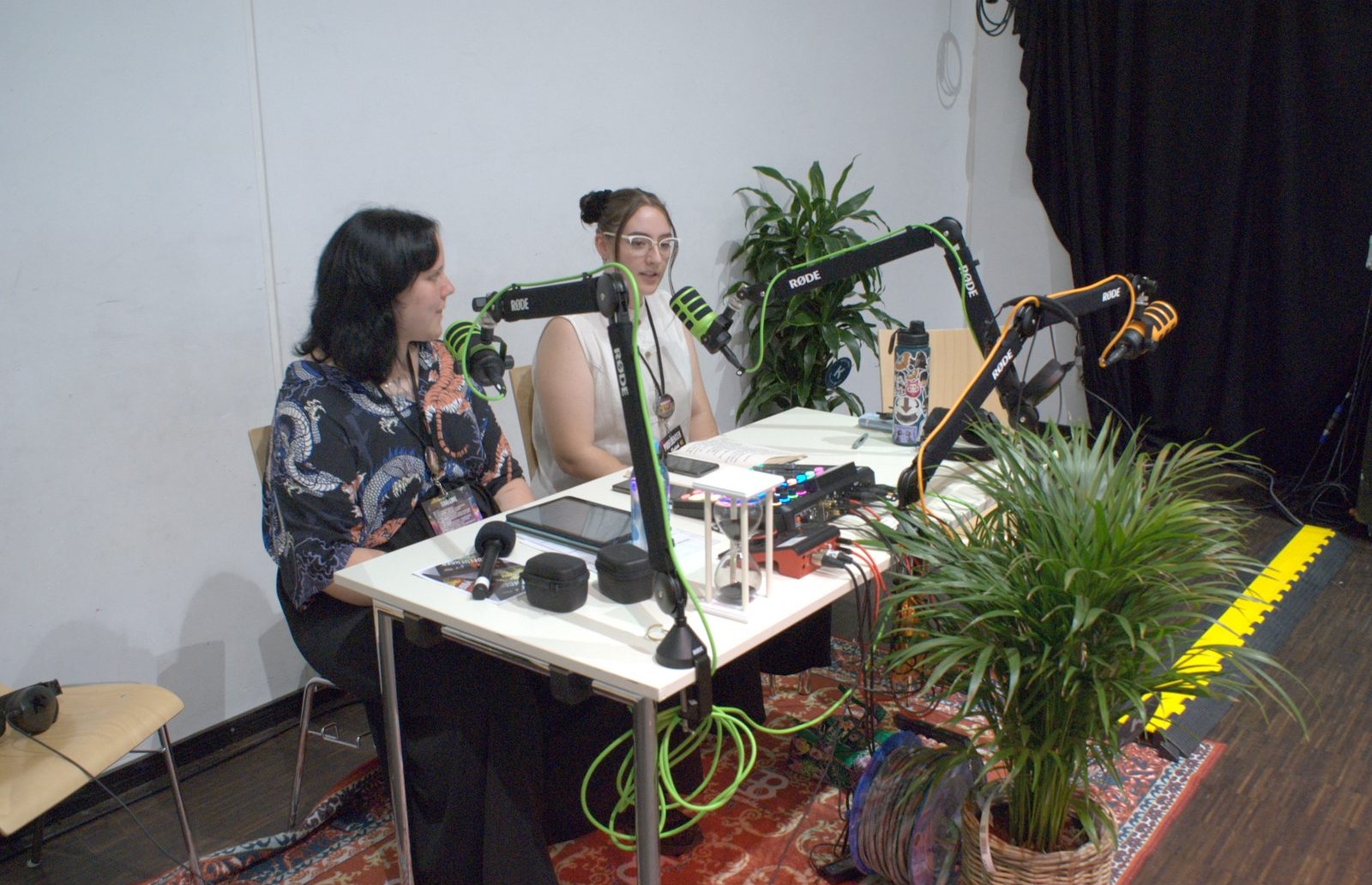 Zwei Podcasterinnen nehmen an einem Studiotisch mit Mikrofonen und Zimmerpflanze eine Folge auf