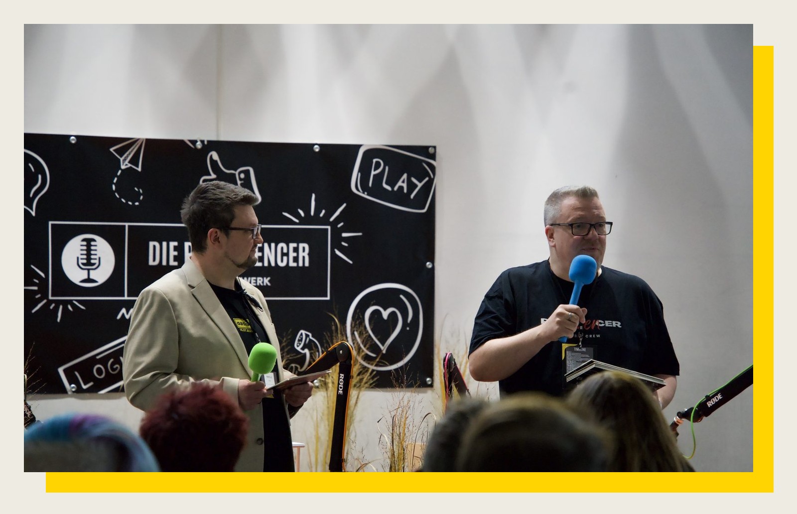 Zwei Moderatoren mit Mikrofonen stehen auf der Bühne vor der Podfluencer-Wand beim Festival