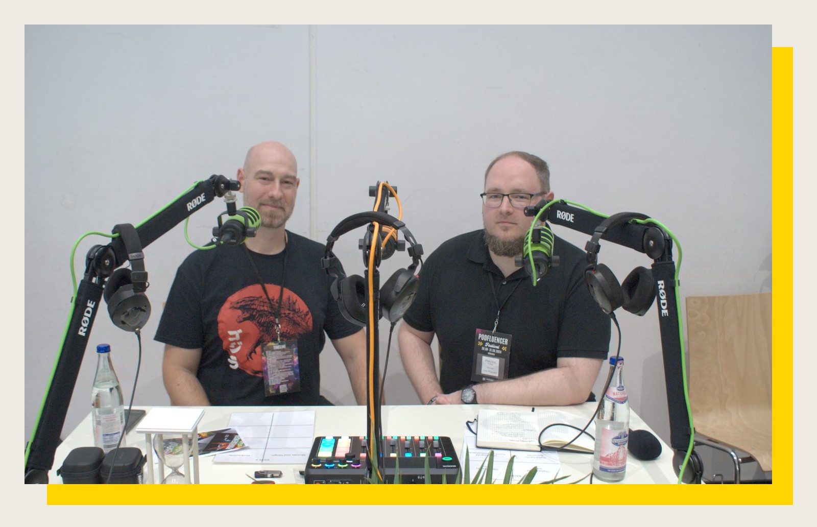 Zwei Podcaster sitzen mit Kopfhörern und Mikrofonarmen am Aufnahmetisch und schauen in die Kamera