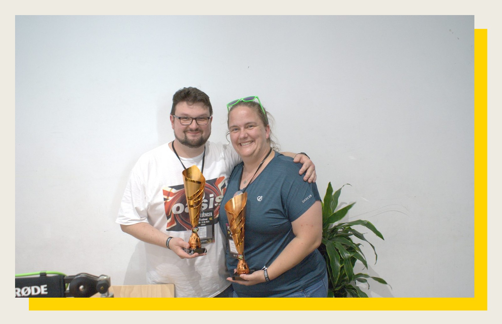 Ein Mann und eine Frau halten lachend ihre Podfluencer-Award-Trophäen vor weißer Wand